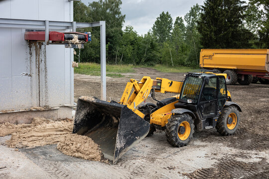 A yellow telehandler cleans up production waste at a high-quality potato starch factory. The heavy machinery scoops wet pulp residue from the ground next to an industrial building.