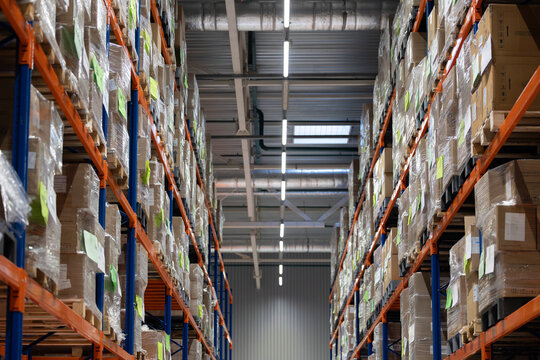 A low-angle view of a tall aisle in a modern logistics warehouse. Industrial orange and blue racks are filled with cardboard boxes on pallets, wrapped in plastic and labeled for shipping.