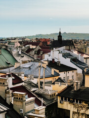 Historic Krakow cityscape revealing weathered rooftops, terracotta tiles, brick chimneys, distant church spire rising against overcast sky, showcasing architectural layering. Elevated view of rooftops