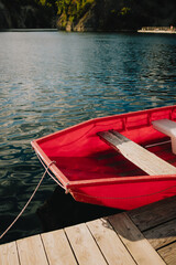 Fototapeta premium Red rowboat with wooden seats gently rocking on calm lake waters, secured to wooden dock, creating serene and inviting atmosphere perfect for relaxation and leisure. Wooden red rowboat floating
