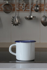 White enamel mug with a blue rim standing on a modern kitchen countertop, featuring a tiled backsplash and blurred hanging cooking utensils in the background creating an appealing still life