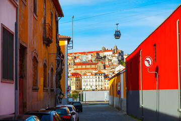 Narrow street view in Vila Nova de Gaia, Portugal, with colorful buildings framing view across Douro River to historic city of Porto. Cable car cabin is visible overhead, ascending towards the hill