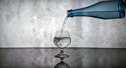 A blue bottle pouring clear water into a stemmed glass on a reflective surface against a gray backdrop