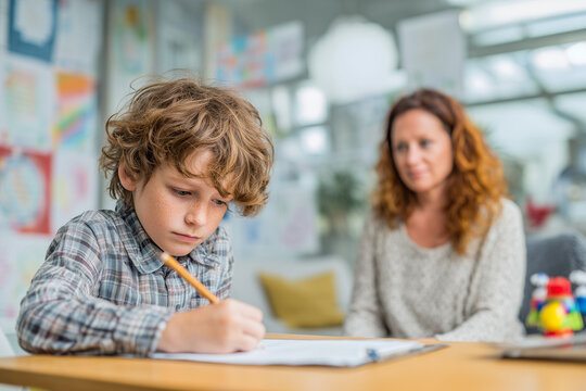 Focused schoolboy concentrating on homework at a desk while a caring teacher watches supportively in a bright classroom — tutoring and learning moment - Powered by Adobe