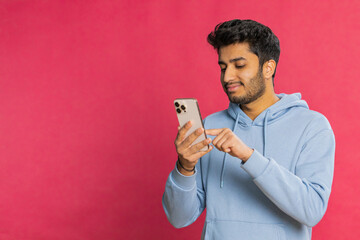 Happy Indian young man browsing social media, smiling while looking at smartphone, enjoying interesting content and typing a message. Relaxed Arabian guy engaging online, isolated on red background