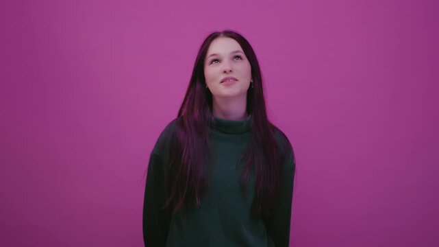 Close up portrait: Caucasian teenage girl in a pink studio in green sweatshirt is lost in thought, looking up. Young brunette woman reflects, creates thoughts. Beautiful female, generation Z or alpha