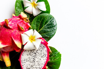 Pitahaya dragon fruits - whole and a half - with frangipani flower and green leaves on white table. Tropical fruits food, top view
