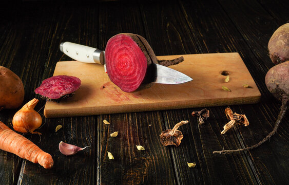 A vibrant beetroot is sliced on a wooden cutting board, surrounded by onions, carrots, and dried spices. The setting has a warm, rustic feel, ideal for cooking
