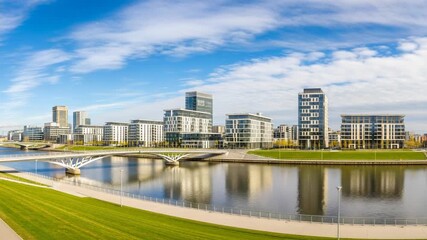 Modern cityscape panorama featuring contemporary office buildings reflected in a wide river under a bright blue sky with scattered clouds
