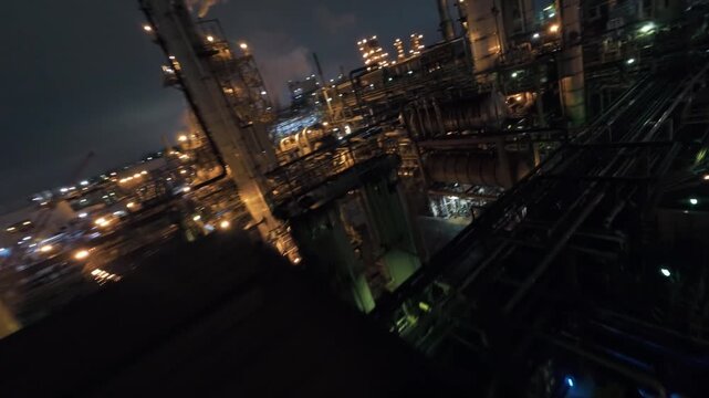 FPV drone dives into glowing energy operation refinery stacks, pipes and tanks as smoke billows into night sky.