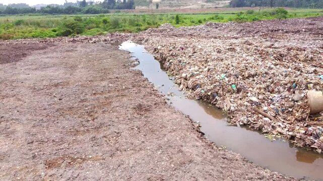 Close-up shot of an enormous landfill waste pile with a channel of dark, polluted leachate liquid runoff flowing beside it, highlighting severe water contamination.
