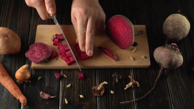 A person is cutting vibrant beets and other fresh vegetables on a wooden board. Garlic and spices are scattered around, highlighting a busy and inviting kitchen environment.