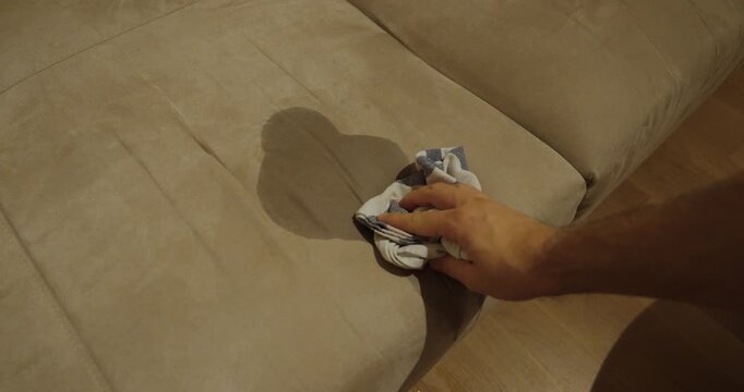 A person gently dabs a stain on a beige fabric sofa with a cloth to remove dirt or liquid.