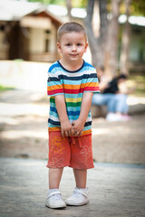 Fototapeta premium A young boy stands in a park wearing a striped shirt and colorful shorts. Friends can be seen nearby, enjoying their playtime in the warm sunshine under trees.