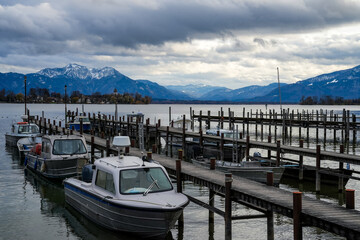 Boats tied to wooden piers on the calm Chiemsee lake with cloudy sky and Alps in autumn