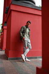 A man in a green jacket stands casually against a bright red wall. Relaxed posture, contrasting colors, and clean composition emphasize a modern casual look.