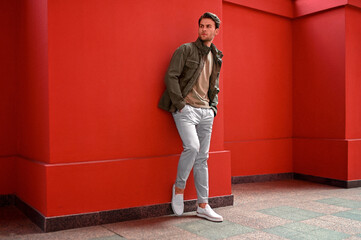 A man in a light brown jacket and white pants walks along a red wall. The clean geometry and muted tones create a minimalist editorial feel.