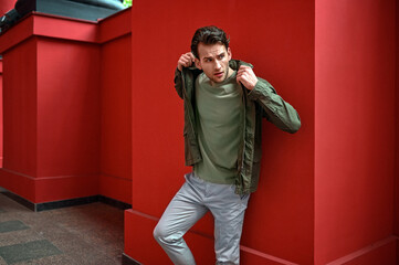 A man in a green T-shirt and lightweight jacket poses by a red wall. Strong color contrast creates a bold, modern street-style look.