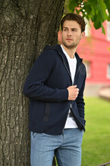 A man in a navy hoodie stands beside a tree. Soft natural light, outdoor greenery, and a relaxed posture create a calm, approachable casual look.