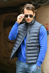A fashionable man adjusts his sunglasses while wearing a gray padded vest over a bright blue shirt. The close-up shot highlights confidence and clean contemporary menswear style.