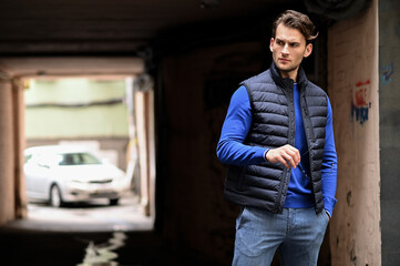 A man in a padded vest and blue long-sleeve shirt stands in a shaded urban area, looking to the side. The contrast of light and dark adds mood while emphasizing the sporty casual outfit.