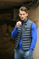 A stylish man in a blue long-sleeve top and padded black vest stands confidently outdoors, holding sunglasses. The portrait highlights casual modern fashion with clean tones and a sharp masculine look