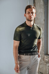 A man in a green T-shirt poses against a concrete wall, standing casually with one knee bent. The raw texture of the background adds a bold, contemporary fashion edge.