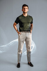 A man in a green T-shirt poses against a concrete wall, standing casually with one knee bent. The raw texture of the background adds a bold, contemporary fashion edge.