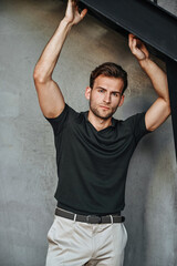 A man in a green T-shirt poses against a concrete wall, standing casually with one knee bent. The raw texture of the background adds a bold, contemporary fashion edge.