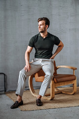 A man in a green T-shirt poses against a concrete wall, standing casually with one knee bent. The raw texture of the background adds a bold, contemporary fashion edge.