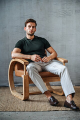 A man in a green T-shirt poses against a concrete wall, standing casually with one knee bent. The raw texture of the background adds a bold, contemporary fashion edge.