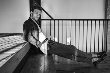 A black-and-white shot of a man relaxing on modern stairs, leaning back with a composed expression. The strong contrast and geometric lines create a cinematic and artistic atmosphere.