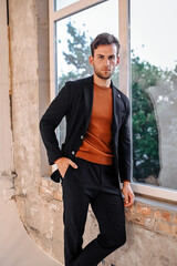 A stylish man in a black blazer and striking orange sweater poses outdoors with a confident expression. The natural backdrop and soft lighting add depth to the modern lifestyle look.
