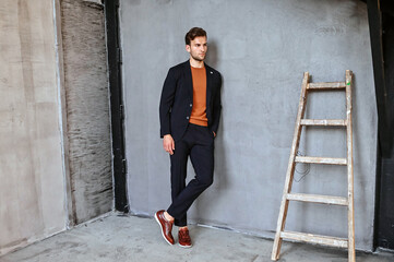 A man dressed in a dark jacket and vibrant orange sweater stands casually beside a wooden ladder in a minimalist industrial space, creating a stylish contrast of textures and colors.