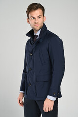 A close-up view of a man wearing a dark navy jacket with his hands casually placed in the pockets. The clean, minimal studio background highlights the refined texture and modern outerwear style.