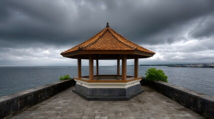 Obraz premium A traditional hexagonal pavilion with a tiled roof stands on a stone platform by the sea under dramatic stormy clouds