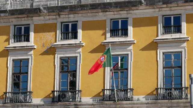 Portuguese national flag waving on the facade of a yellow building under bright sunlight in Lisbon, Portugal.
