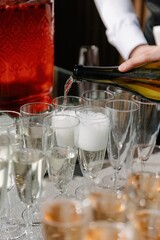 A bartender pours sparkling wine from a bottle into several tall champagne flutes, creating bubbles, on a catering table with a red drink dispenser nearby.