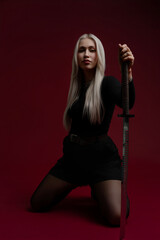 Woman in black outfit poses with sword against a red background in a dramatic setting