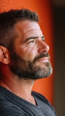 Fototapeta premium Man with a salt and pepper beard looks upward and to the right. He is wearing a dark shirt and is posed against a bright orange wall