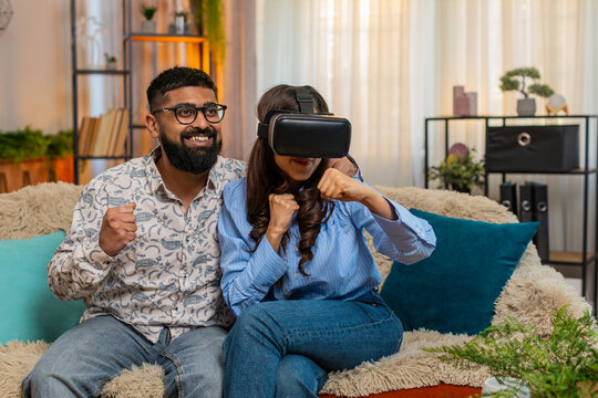 Indian couple man woman sitting on home sofa, girl wearing VR headset laughing while boy supports her excitement during virtual game. Young family enjoying funny immersive experience indoors together - Powered by Adobe