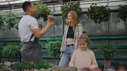 A male sales consultant gives advice for buying plants for mother and daughter. A male plant botanist communicates with a visitor to a plant store. Woman wants to buy a green plant in a store