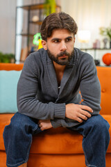 Fototapeta premium Indian man at home bends forward holds head in hands strong fatigue slow breath eyes down seeks rest. Arabian guy on sofa sits still rubs temples tries to calm mind seeks relief from stress and pain.