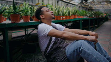Man resting on greenhouse floor surrounded by potted plants in a serene environment after a long day of work