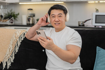 Asian man at home wears wireless headphones listens to playlist relaxes closes eyes and smiles. Chinese guy on sofa enjoys calm music breathes slow feels peace rests and recharges mood with soft sway.