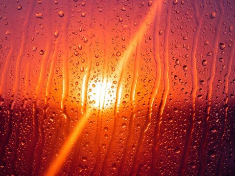 Raindrops and streaks cover a windowpane, viewed against a vibrant orange and red sunset or sunrise, creating a dramatic, glowing abstract effect.