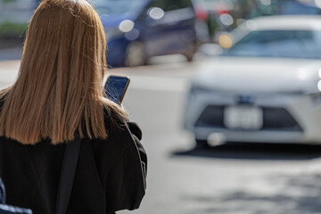 Young woman with blonde hair stands on a sunny urban street texting on smartphone as she waits to cross a street where blurred cars pass in the background.