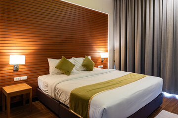 A bed with white sheets and green pillows sits against a wooden panel wall, with two wall lamps and small side tables creating a simple layout.