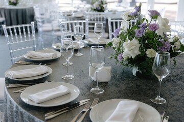 Elegant wedding reception table set with white plates, fine cutlery, crystal glasses, and a large floral centerpiece of white roses and purple eustoma