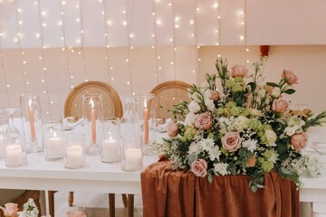 Romantic wedding table setting featuring a lush floral arrangement with blush roses, various lit white and salmon candles in glass holders, and a background of warm fairy lights